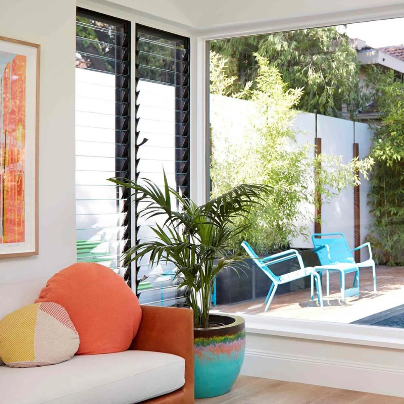Living room with a couch, colorful pillows, and a large louvre window overlooking a garden.
