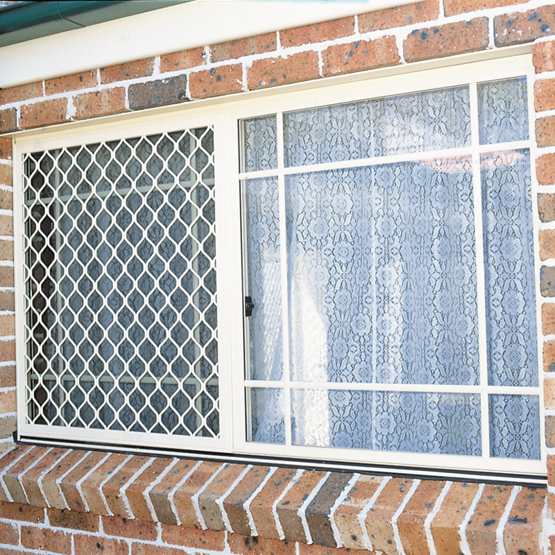 A white-framed sliding window with a diamond-pattern security screen on the left panel and lace curtains inside, installed on a red brick wall.