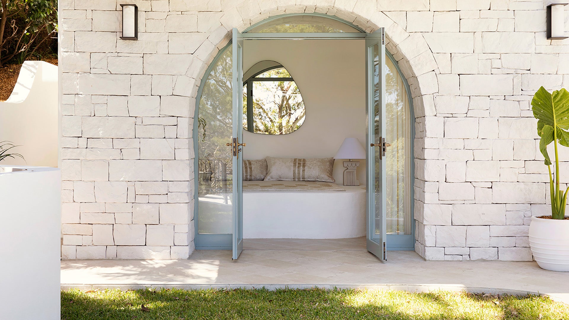 Open timber hinged door leading to a sunlit room with white walls and a bed, framed by a stone wall.