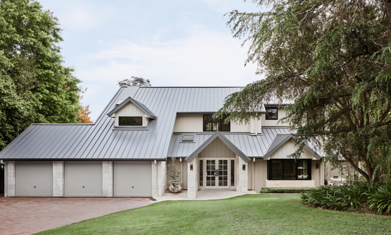 A modern two-story house with a gray metal roof, beige siding, a three-car garage, and a landscaped lawn with trees.