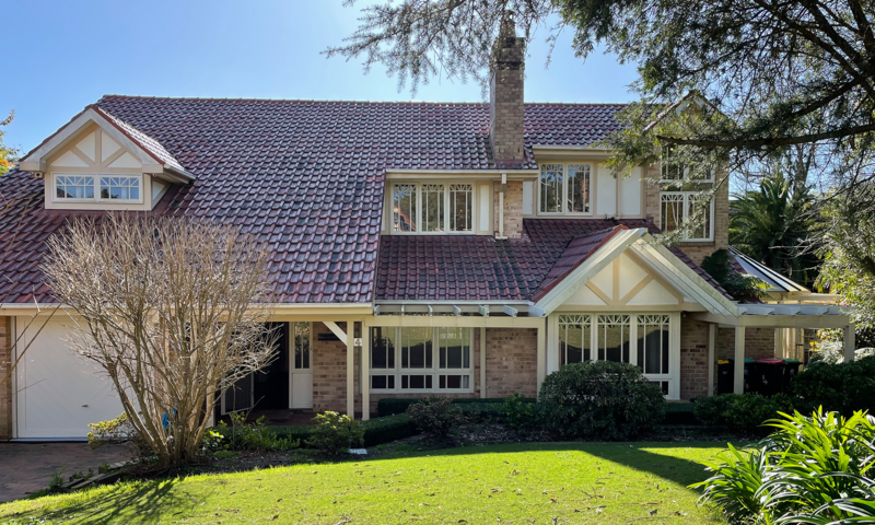 A large residential house with a dark red tiled roof, cream and brick exterior walls, multiple windows, a garage on the left, and a green lawn in front under a blue sky.