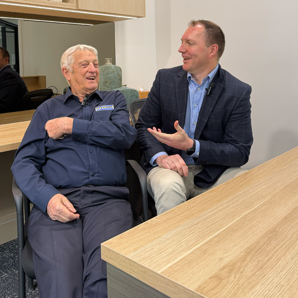 Two men sitting and talking in a modern office or meeting space. The man on the left wears a navy blue long-sleeve button-up shirt and dark trousers, while the man on the right wears a navy check blazer over a light blue dress shirt and khaki pants.